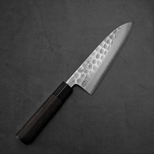The Yoshikane tsuchime SKD santoku 165mm knife by Yoshikane, featuring a hammered SKD steel blade and a dark wooden handle, is placed diagonally on a gray textured surface.